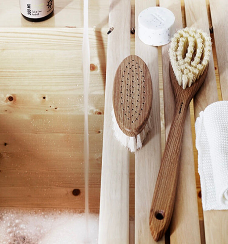 A bath brush with a wooden handle and white bristles and one without a handle, laying on a wooden surface with a bamboo bath mat and a wooden sponge in the background. A bottle of bath salt is also visible in the top left corner.
