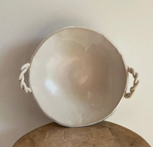 Round beige ceramic bowl with rope handles on a wooden table against a plain background