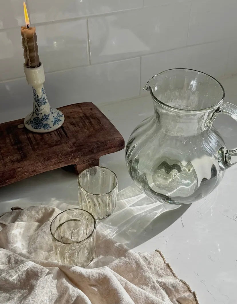 Clear glass pitcher and candle holder on a white surface with a wooden riser and two glasses.