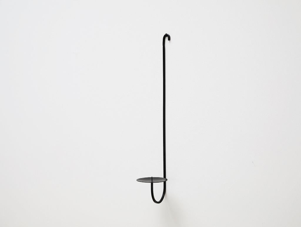 A minimalist black iron candle holder with a single arm, mounted on a wall, with a small platform for holding a candle.