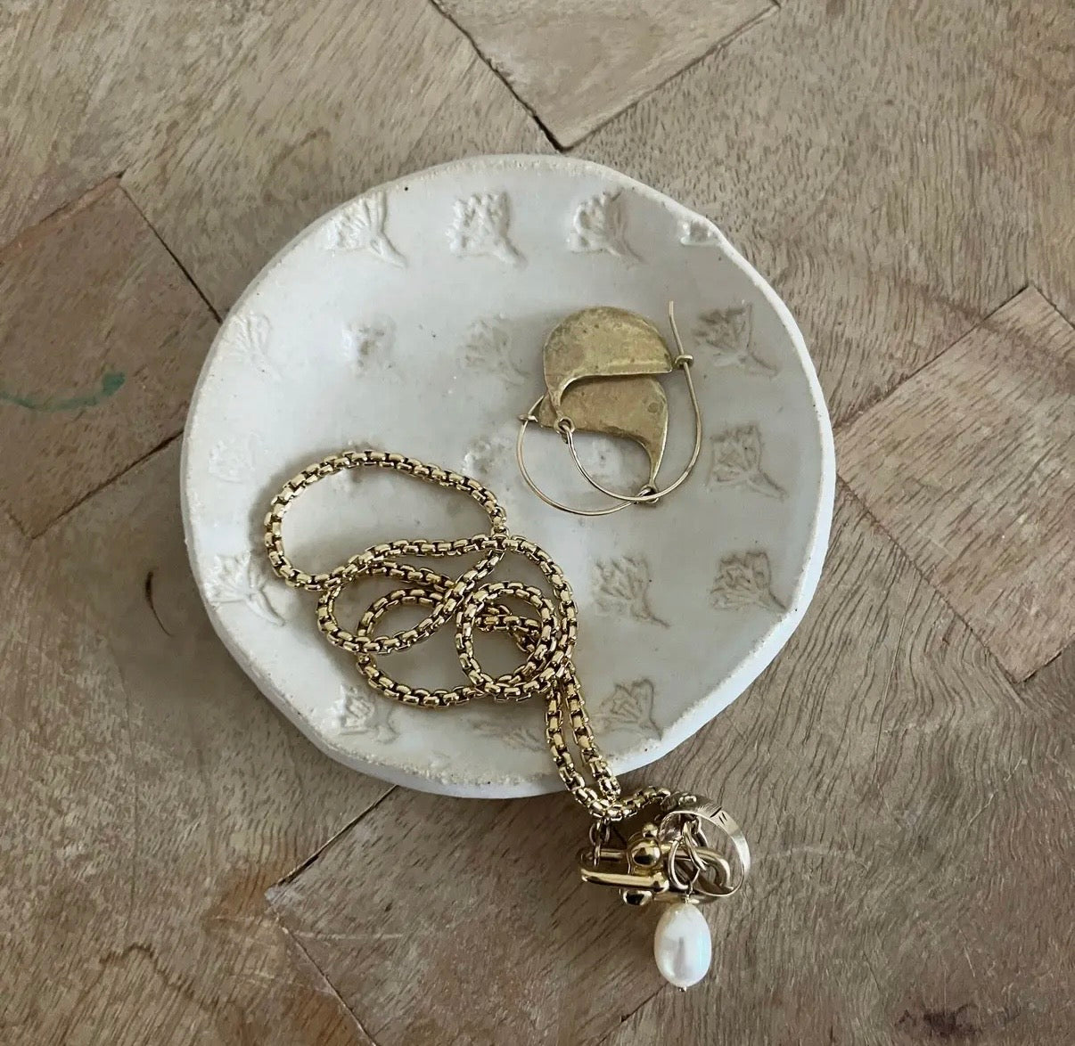 Gold necklace and hoop earrings on a textured white dish on a wooden floor.