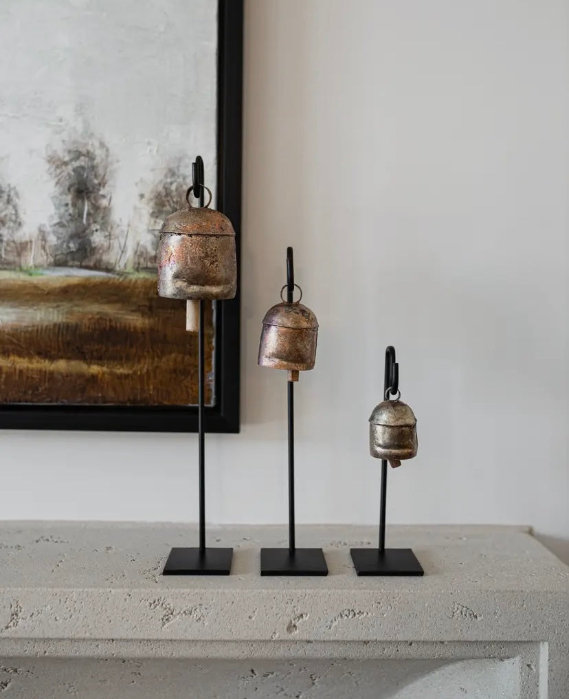 A set of three copper bells of varying sizes, each mounted on a welded iron hook, displayed on a mantle.