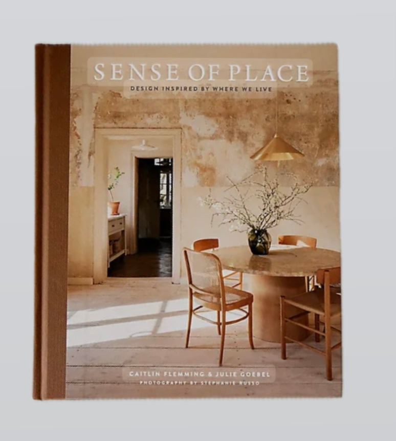 The cover of the book 'Sense of Place' by Caitlin Flemming and Julie Goebel, featuring an interior shot of a home with a wooden dining table and chairs, leading to a hallway with a soft view of a room with a bookshelf.