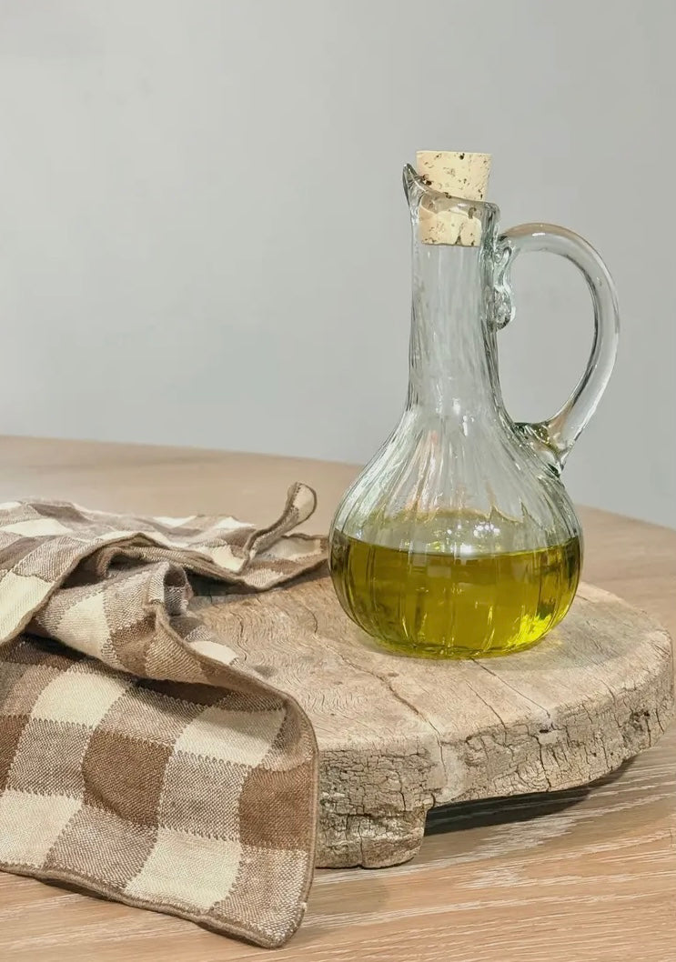 Glass cruet with cork stopper containing oil on a wooden board with a checkered cloth.