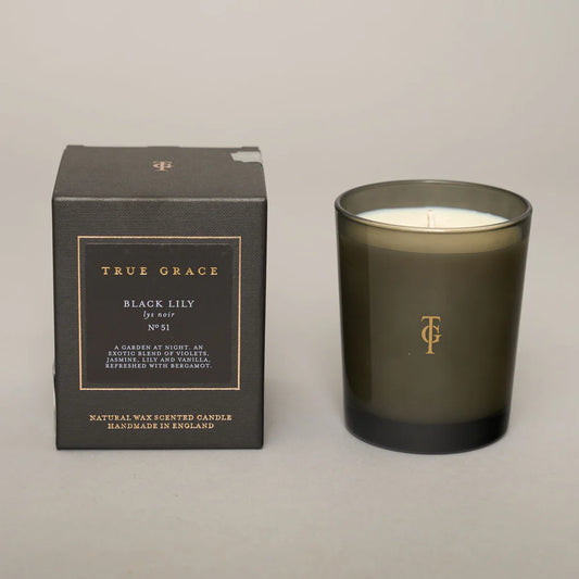 A black candle with a label that reads 'Black Lily No.51' and the brand 'True Grace' is displayed next to its black box packaging with the same text.