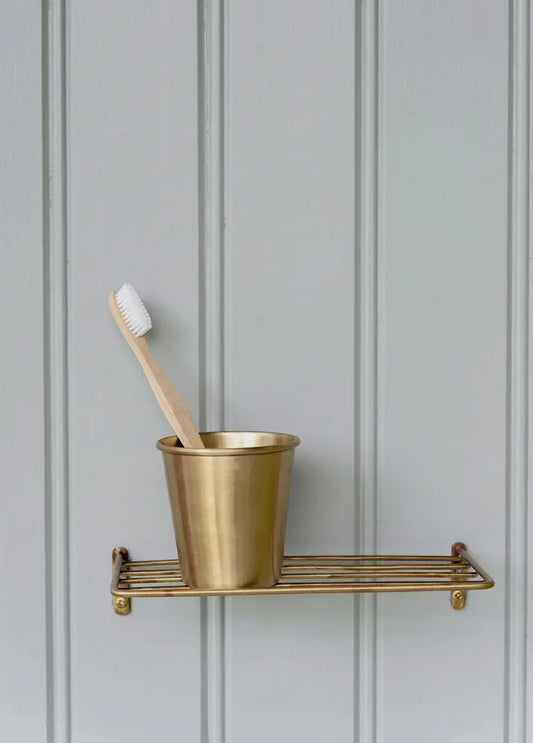 A brass metal tumbler placed on a matching metal wire shelf, against a grey panel background, with a toothbrush inside the tumbler.