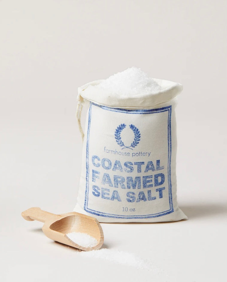 A paper bag labeled 'COASTAL FARMED SEA SALT' with a wooden spoon in front, indicating the product is a natural sea salt harvested from East Coast waters.  By Farmhouse Pottery.