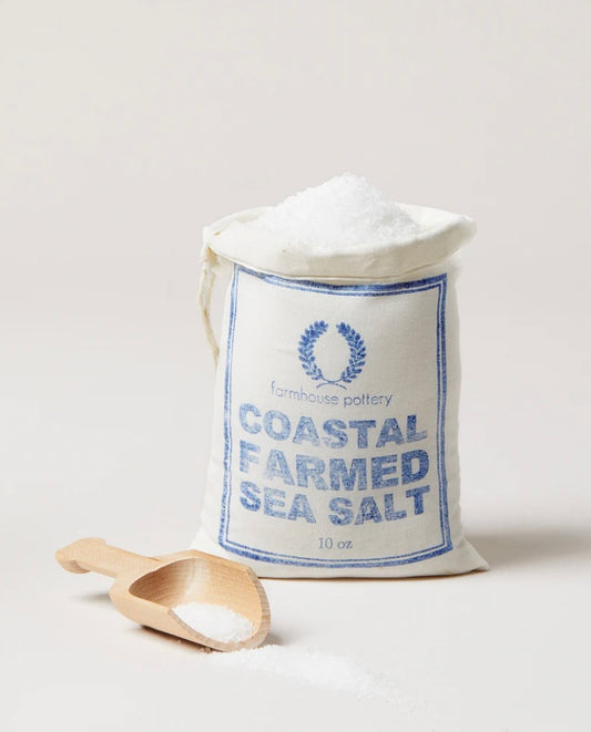 A paper bag labeled 'COASTAL FARMED SEA SALT' with a wooden spoon in front, indicating the product is a natural sea salt harvested from East Coast waters.  By Farmhouse Pottery.