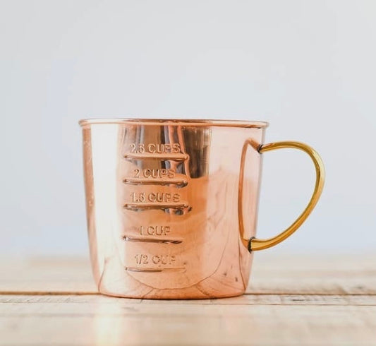 A copper measuring cup with interior and exterior markings and a brass handle, displayed against a light background.
