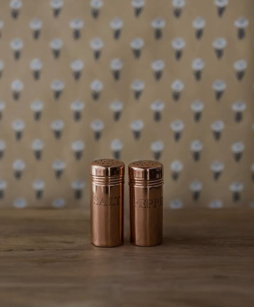 Two bronze salt and pepper shakers on a wooden surface with a patterned background.