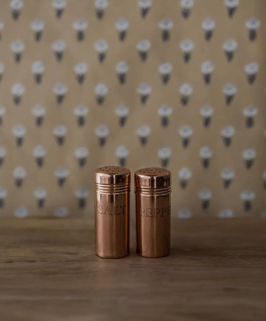 Two bronze salt and pepper shakers on a wooden surface with a patterned background.