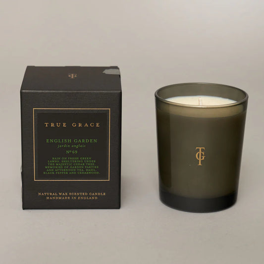 A green glass candle with a black lid and label that reads 'True Grace English Garden Pure Essence'. There is also a black box with 'True Grace' and product details visible in the background.