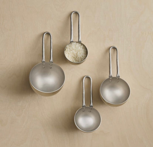 A set of four pewter finish forged measuring cups with handles, displayed on a wooden surface. Each cup has a different size, with one filled with rice to indicate capacity.