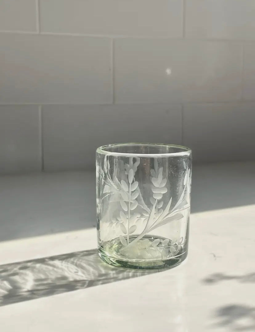 Clear glass tumbler with floral etching on a light surface with a tiled wall background