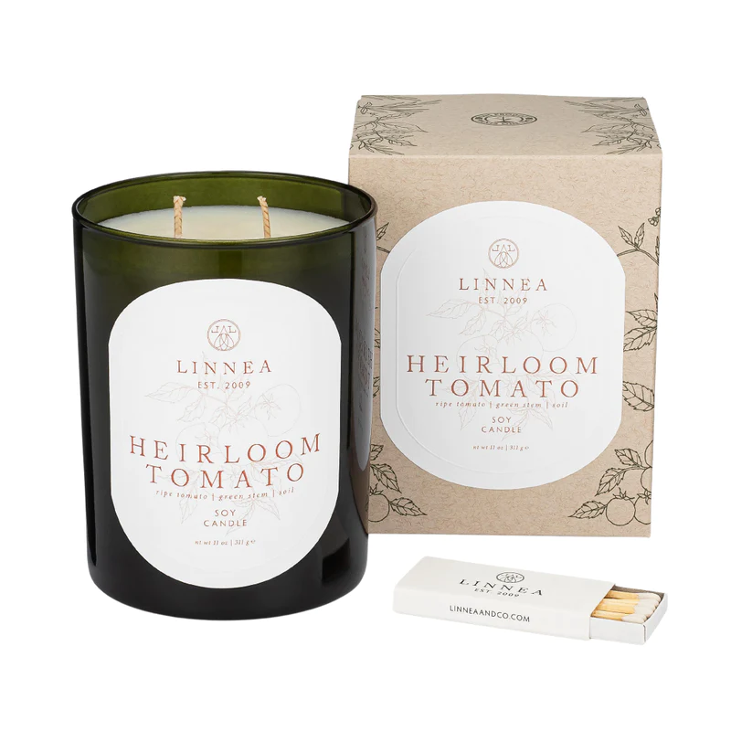 A green Linnea candle with a label that reads 'Heirloom Tomato', alongside a brown box with the same label and an illustration of a tomato plant. A matchbook is also visible.