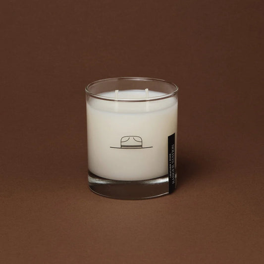 A clear glass candle with a white wax layer, featuring a minimalist design with a line drawing of a hat on the label.