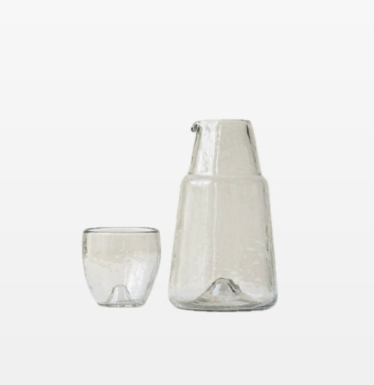 A clear glass bedside carafe set with a tapered design and a rounded glass lid, both items are transparent and displayed against a white background.