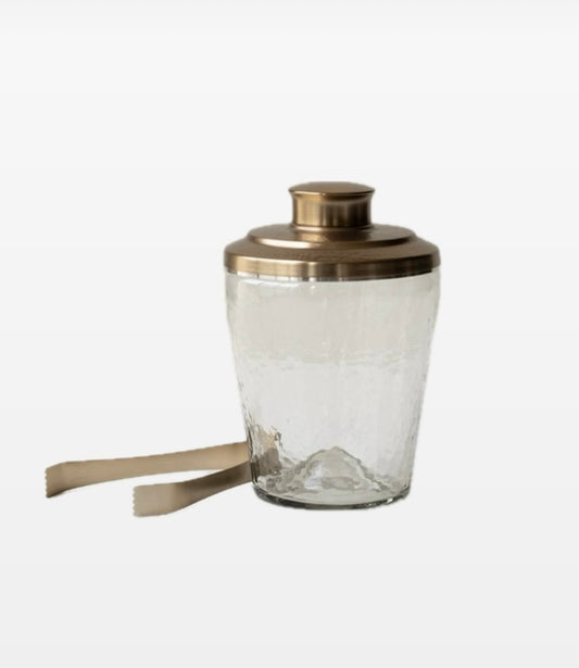 A clear glass ice bucket with a hammered brass lid and a pair of brushed brass tongs, with a beige strap attached to the bucket.