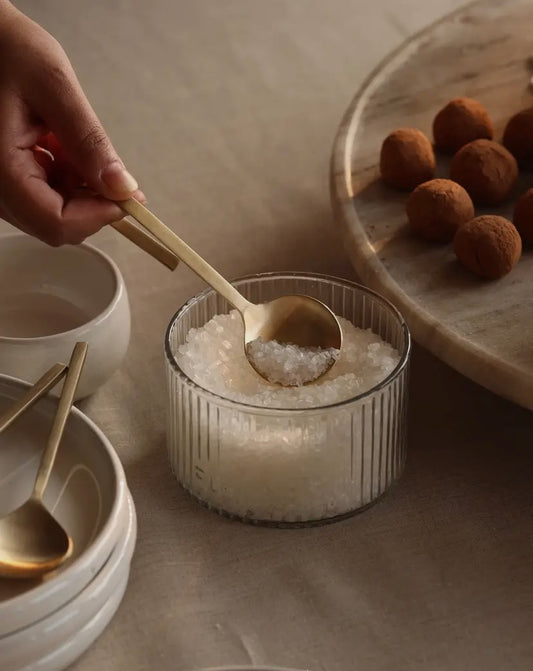 A person's hand is using a brass spoon to scoop salt from a clear container, with the salt visible in the spoon. The setting includes a wooden table and a collection of round white dishes in the background.