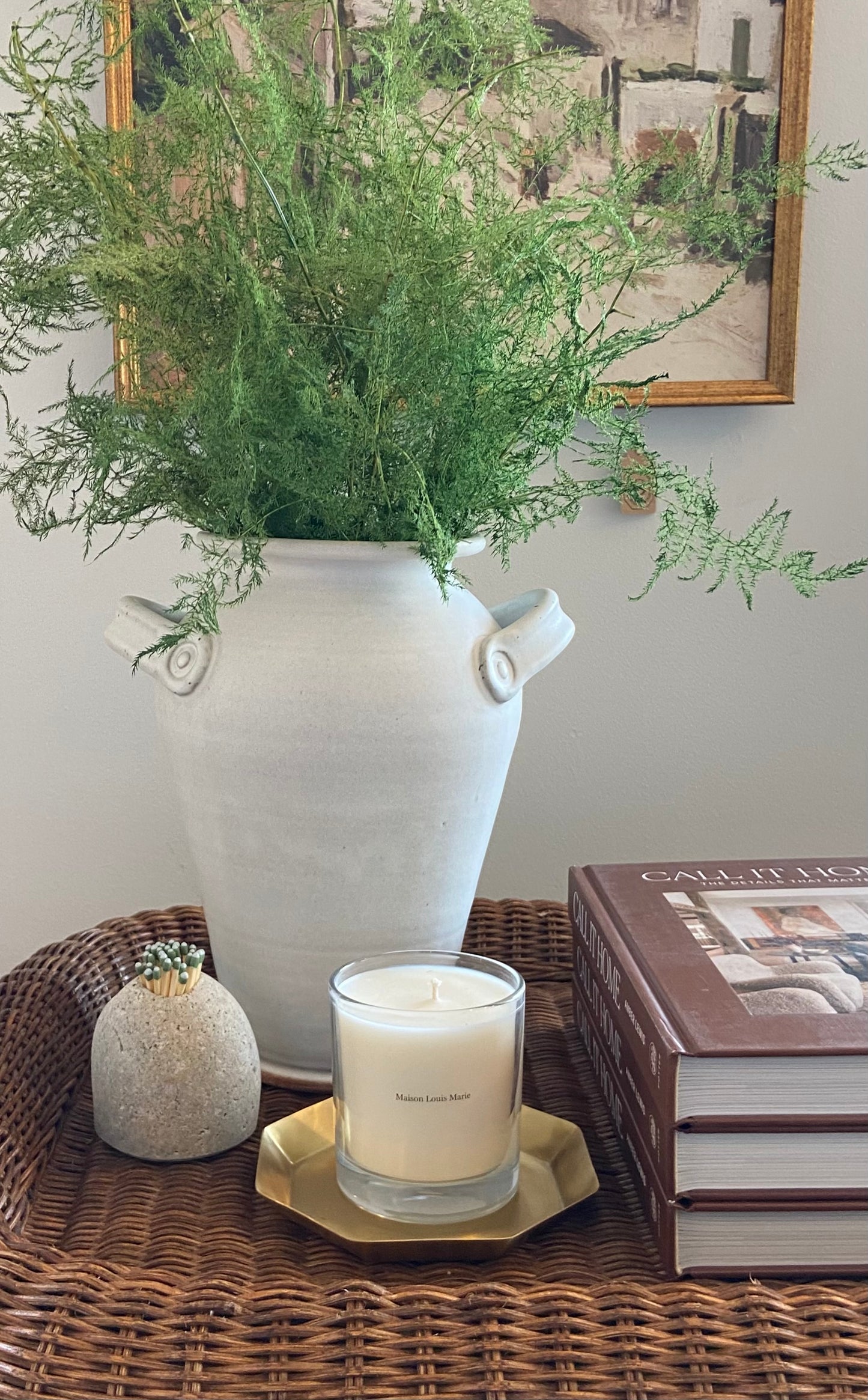 Lyon vase with greenery on a wicker tray with a candle, match striker, and books in the background.