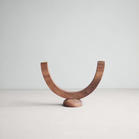 A wooden candelabra with a sculpted crescent shape balanced on a smooth, domed base with two solid brass candle cups.