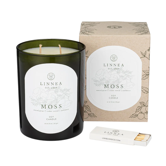 A black jar candle with a white label reading 'MOSS', accompanied by a brown kraft box with floral designs and a match striker.