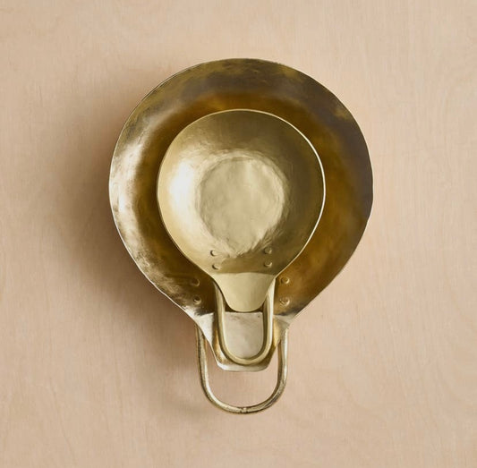 A gold-colored, pan-shaped metal spoon rest with a hammered surface and a handle, displayed against a neutral background.
