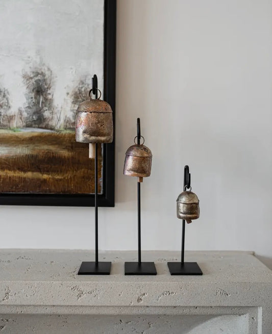A set of three copper bells of varying sizes, each mounted on a welded iron hook, displayed on a mantle.