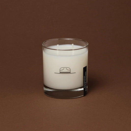 A clear glass candle with a white wax layer, featuring a minimalist design with a line drawing of a hat on the label.