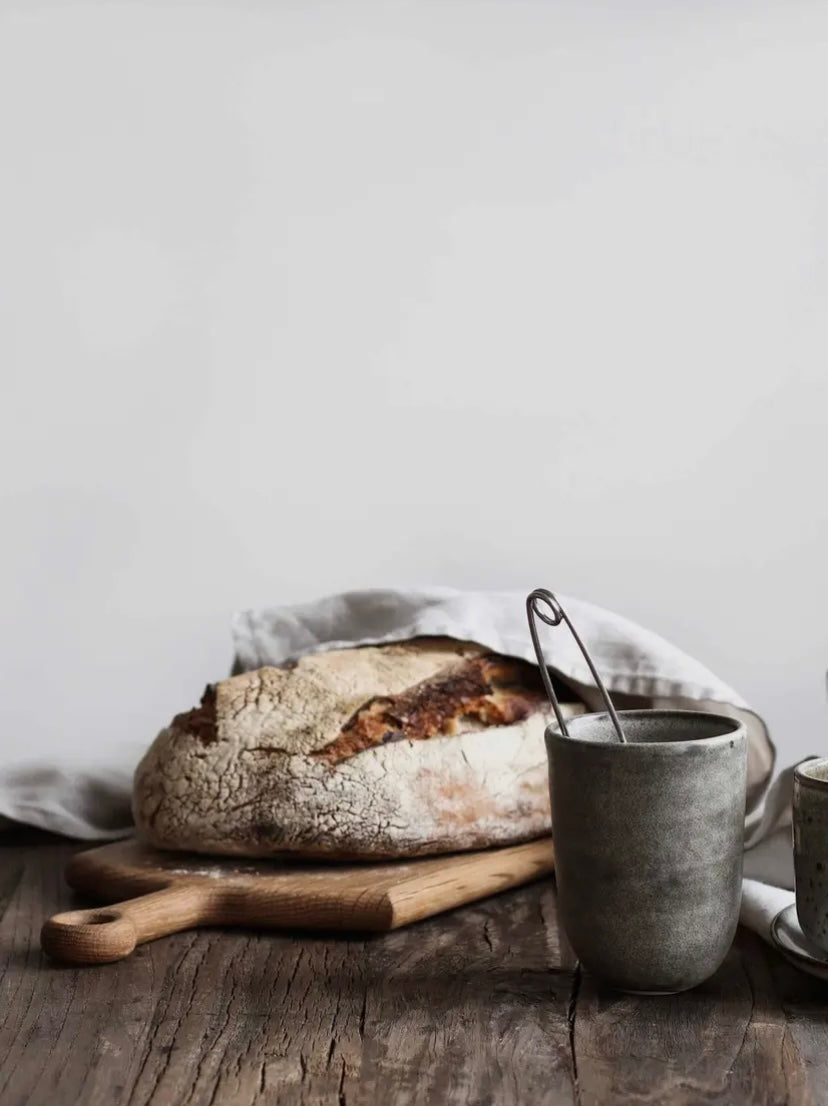Loaf of bread on a wooden board with a ceramic mug on a rustic wooden surface.
