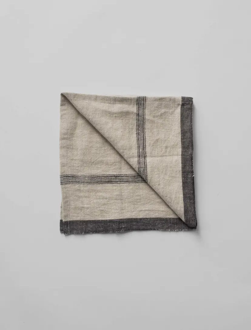 A beige linen napkin with denim colored stripes, displayed on a neutral background.