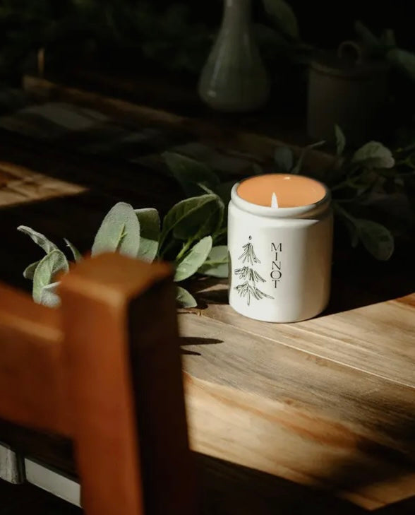 Candle by MINOT in a white container with a tree design on a wooden surface with greenery