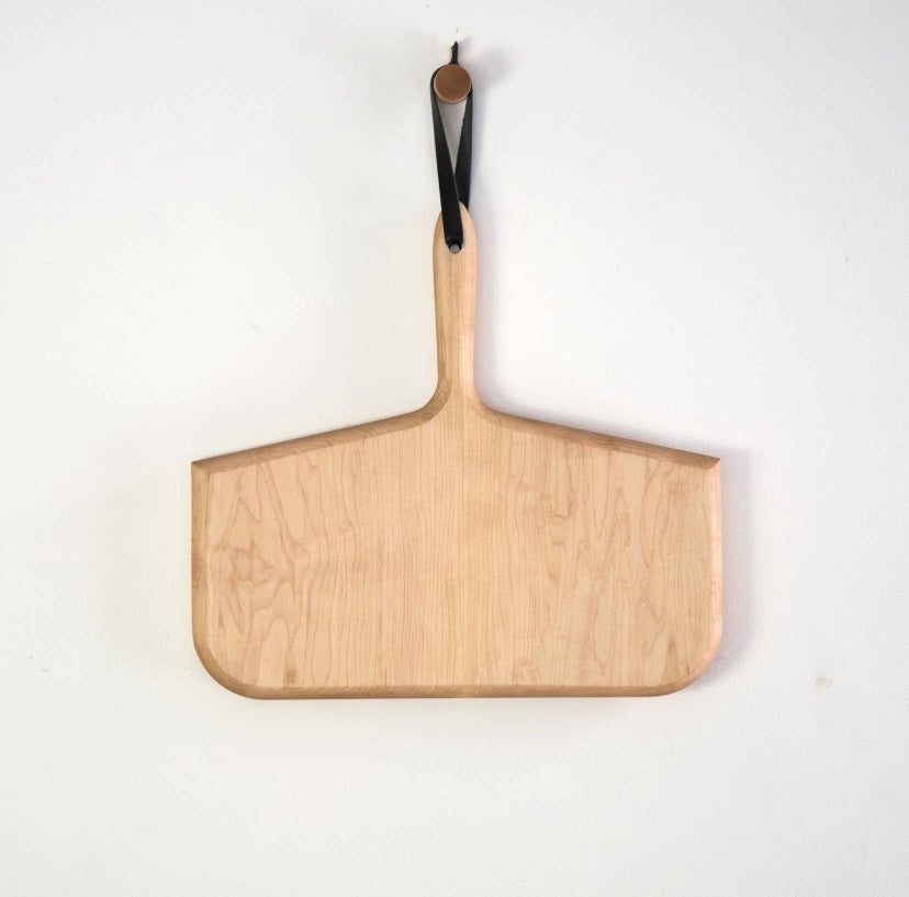 A maple wood cutting board with a balck handle and a hanging loop, displayed against a white background.