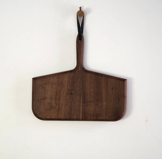 A walnut wood cutting board with a handle, hanging on a white wall.