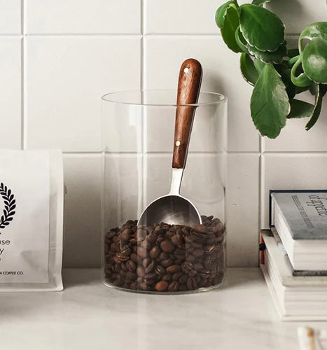 ARTISAN COFFEE SCOOP Farmhouse Pottery in a glass cannister filled with coffee beans on a counter with books off to the side.