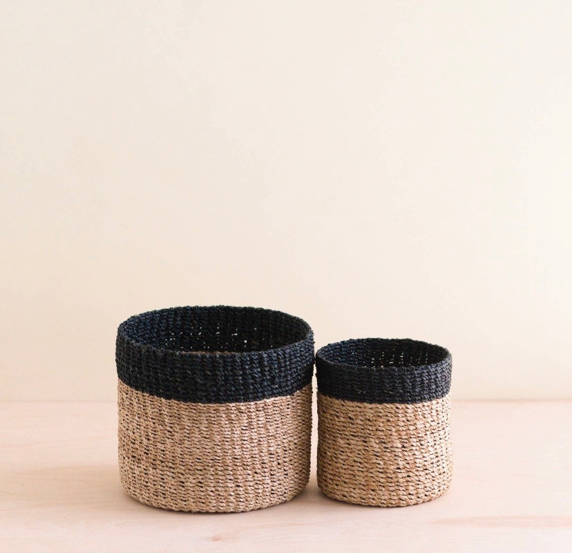 Two handwoven baskets with black and natural colors, one small and one medium in size, placed on a wooden surface against a neutral background.